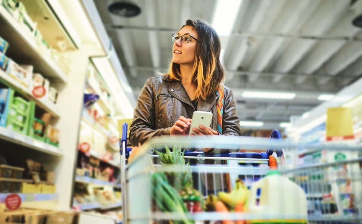 Photo shows a young woman shopping in a supermarket while holding her phone and pushing a trolley